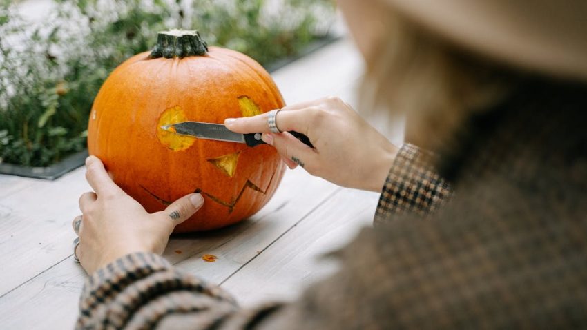 A woman carves a Halloween pumpkin with a knife on a wooden table outdoors.